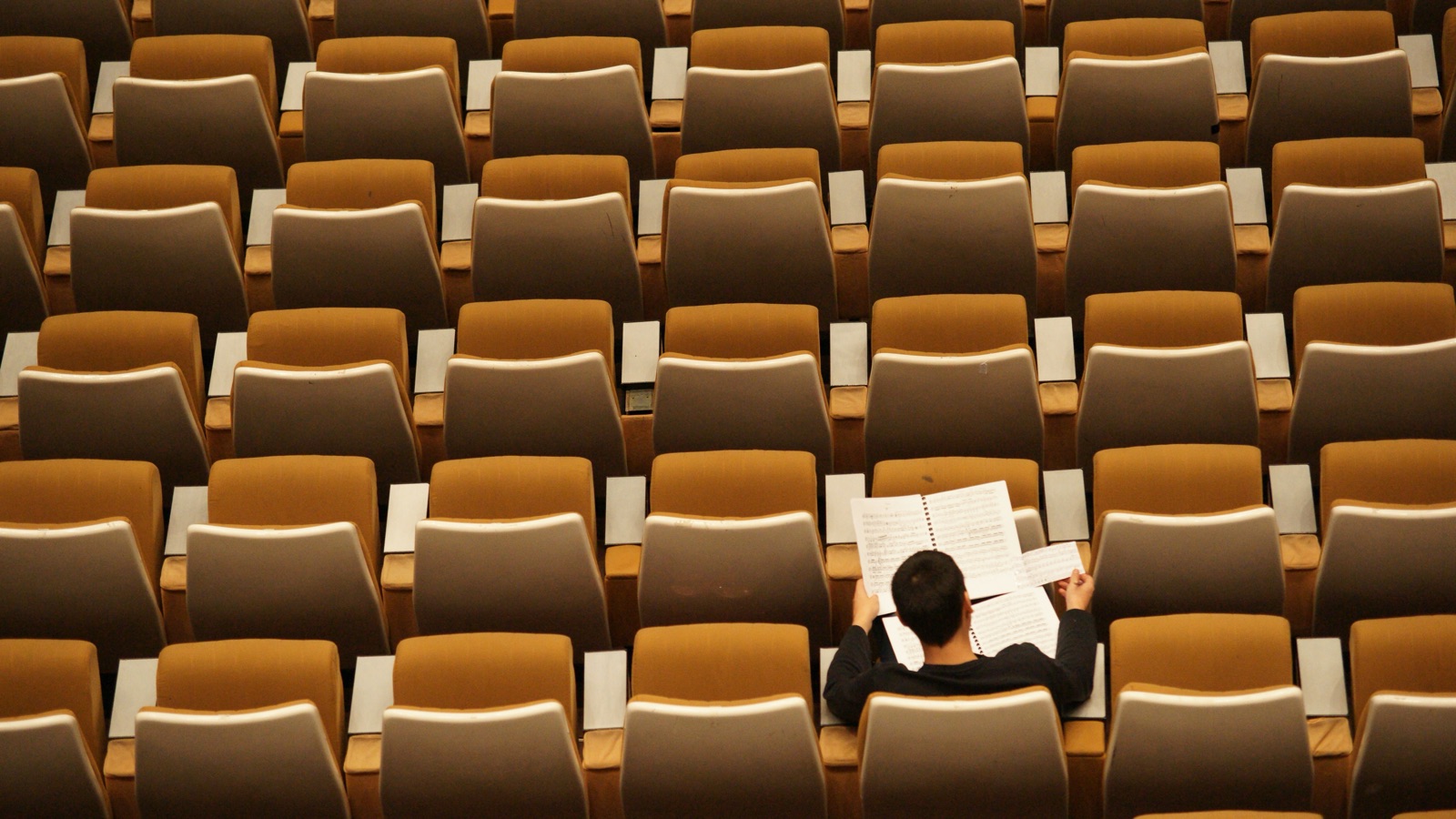 A student sitting alone among rows of empty lecture hall seats, reading through sheet music