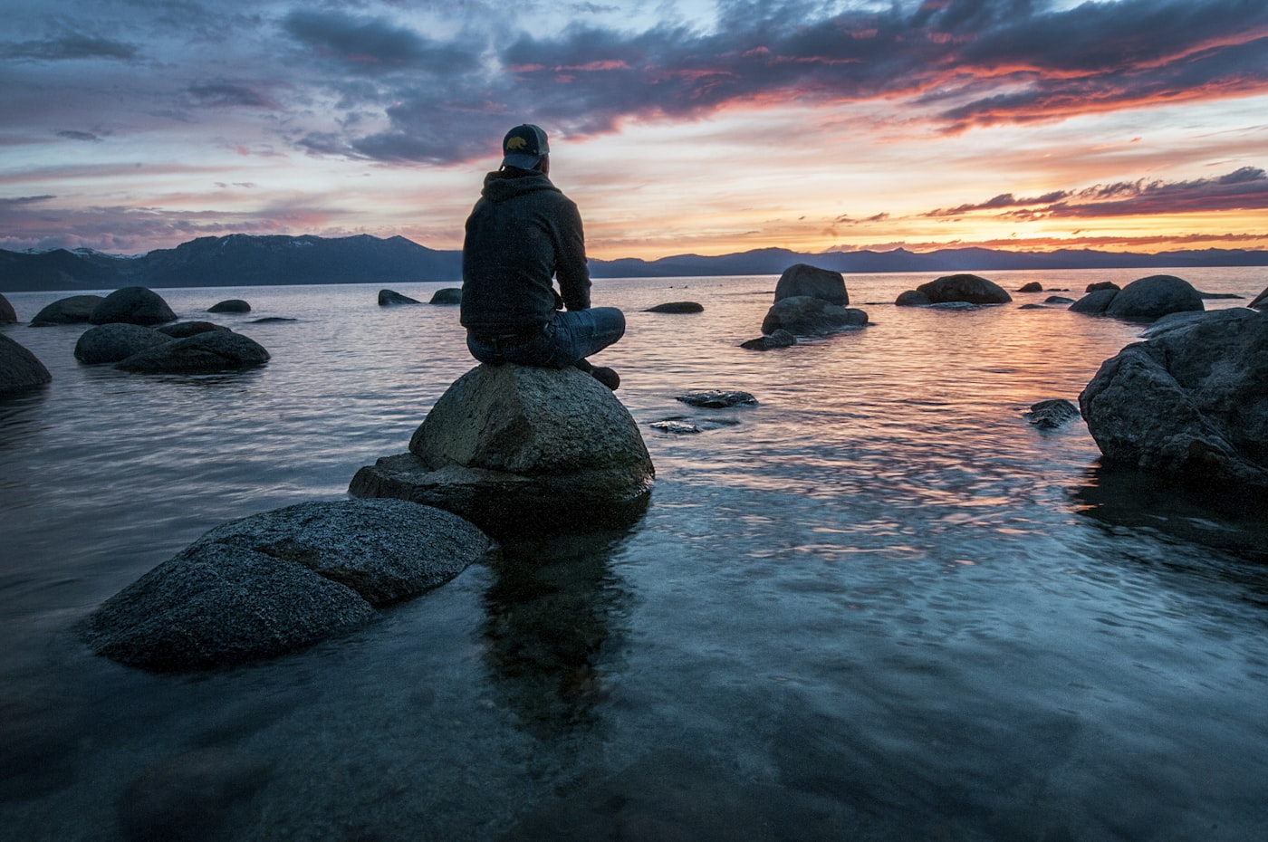 A person sitting in quiet meditation on a rock at the water’s edge at dusk